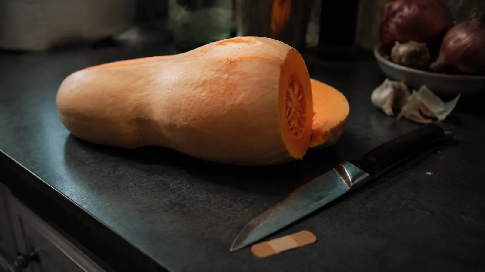 A butternut squash half-cut on a wooden cutting board with a chef's knife resting beside a small bandage