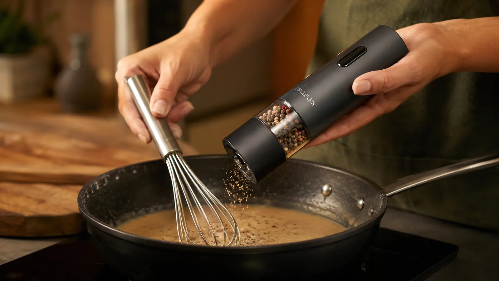 A hand tilting the Circle Joy electric pepper grinder sideways over a pan of gravy while the other hand holds a whisk