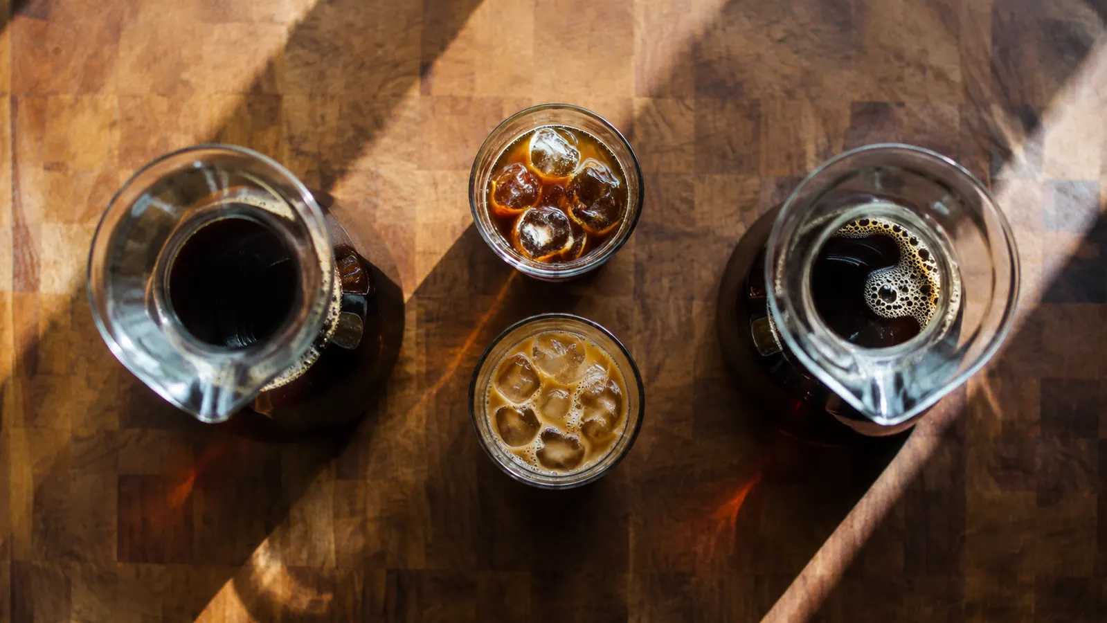 Primula Burke Deluxe cold brew carafe and a Hario Mizudashi pitcher sitting side by side on a kitchen counter with glasses of iced coffee between them